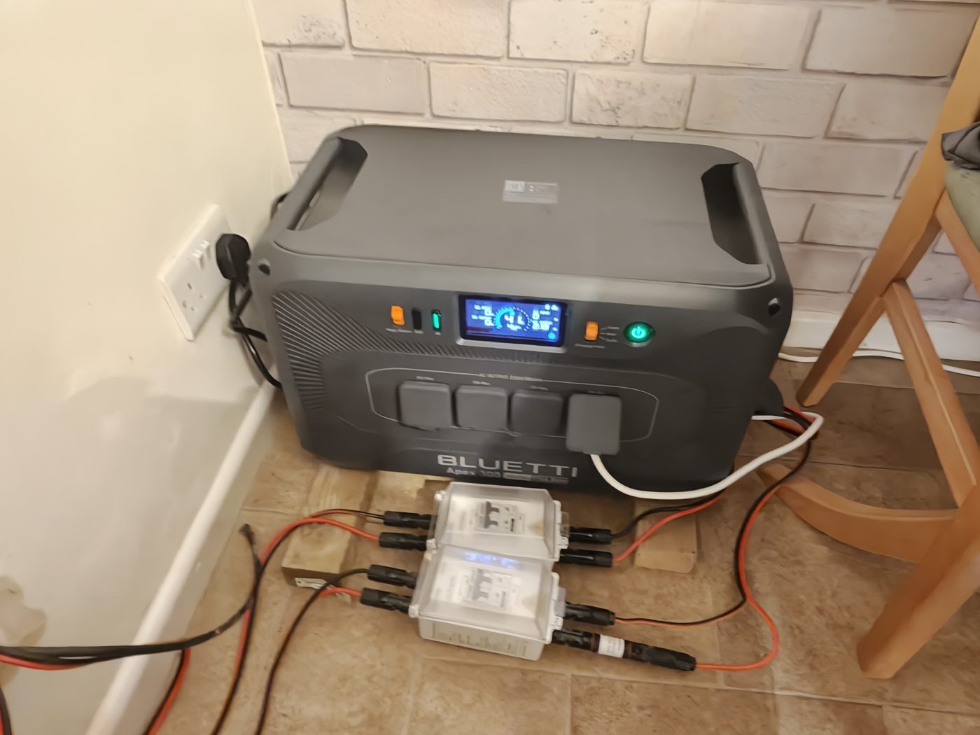 My Bluetti Apex 300 on the floor against a brick wall, plugged into the mains, with red and black solar cables running through two isolator switch boxes in front of it.
