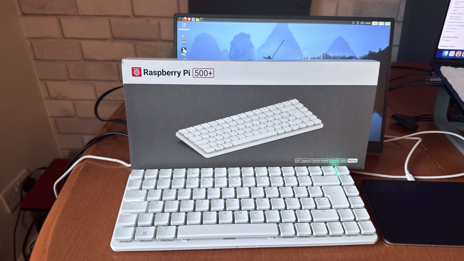 My white Raspberry Pi 500+ keyboard computer on a wooden desk, in front of its box, which shows UK layout, 16GB RAM and 256GB SSD, with the Raspberry Pi desktop on a monitor behind.