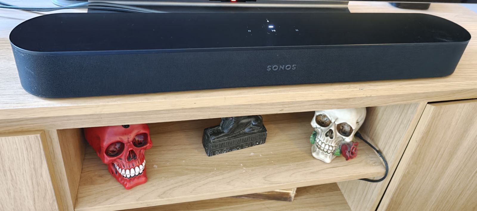 My black Sonos Beam Gen 2 on a wooden TV unit, in front of the TV, with decorative skulls on the shelf below.