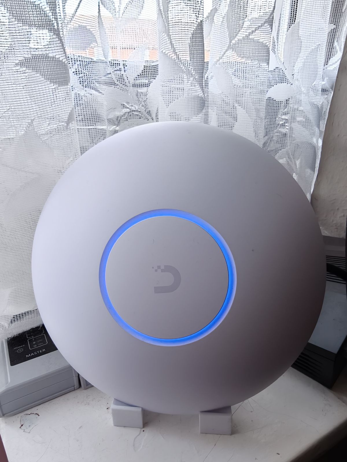 My UniFi U7 Pro access point standing upright on the lounge windowsill on a white 3D-printed stand, with its blue LED ring lit.